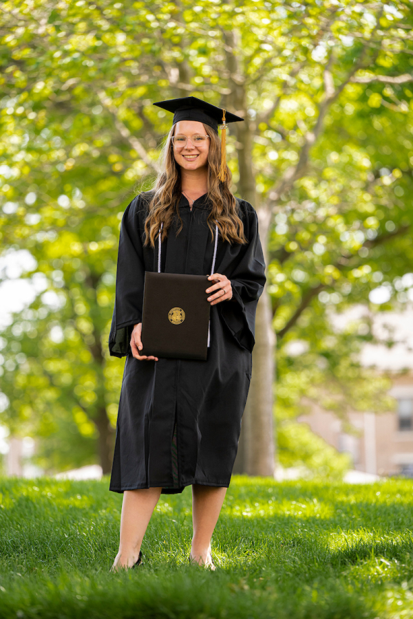 a female graduate student in regalia and holding a diploma pictured outside during spring time